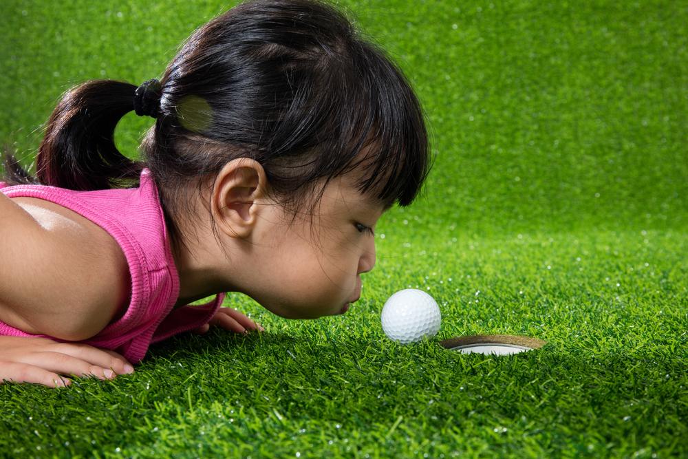 Little asian girl blowing on a golf ball to make it go in the hole.