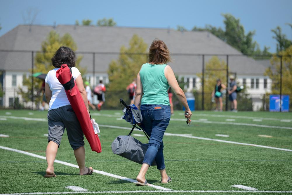 Two moms walking on a field before a youth sports game carrying chairs.