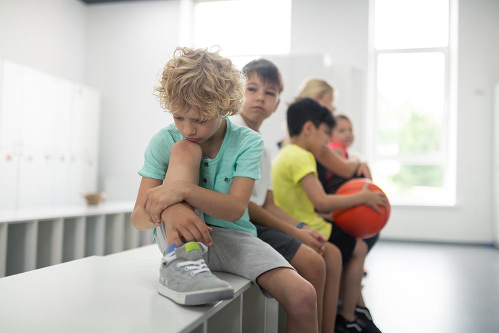 Young white boy sitting next to other boys on a locker room bench looking sad.